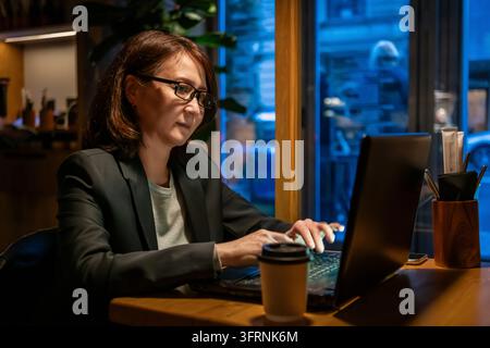 Femme mûre portant des lunettes travaillant sur un ordinateur portable à une table en bois dans un café confortable, éclairée par une lumière intérieure chaude et des reflets bleus de fenêtre. Banque D'Images
