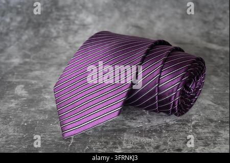 Une cravate classique pour homme en soie violette foncée à fines rayures blanches diagonales, posée sur une surface texturée en ardoise sombre. Accessoire de mode haut de gamme pour homme Banque D'Images
