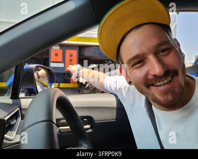 Un homme barbu avec un chapeau jaune sourit et pointe un bouton rouge sur un camion. Il porte une chemise blanche et un chapeau noir Banque D'Images