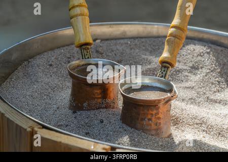 Grossissement de deux petites cezves en cuivre préparant du café dans le sable chaud, montrant une méthode de préparation traditionnelle avec des manches en bois et un métal rustique chaud Banque D'Images