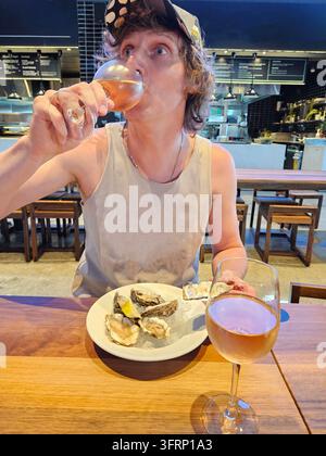 Un homme boit du vin dans un verre. Il porte un débardeur blanc. Une assiette d'huîtres est posée sur une table avec des verres de vin Banque D'Images