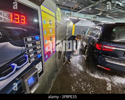 Un homme lave sa voiture lui-même. Le lave-auto a un panneau qui indique 12. Texte - le dispositif de distribution ne distribue pas, eau, osmose, mousse, payer, prendre le pistolet, sélectionner un programme, démarrer le lavage, garder propre Banque D'Images