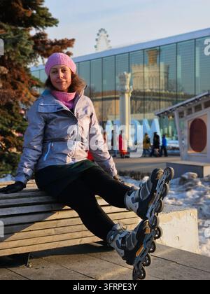 Une femme est assise sur un banc avec ses patins à roulettes à la VDNH. Elle porte un chapeau rose et une écharpe rose. Grosses lettres - ATOM Banque D'Images