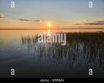 Le soleil se couche sur un plan d'eau calme, projetant une lueur chaude sur la scène. L'eau est immobile et paisible. Le ciel est un mélange de teintes orange et roses. Lac Pleshcheevo à Pereslavl-Zalessky Banque D'Images