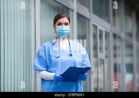 Médecin NHS debout devant un établissement médical portant un uniforme bleu, un masque chirurgical de protection, des gants bleus et un stéthoscope, tenant un patient Banque D'Images