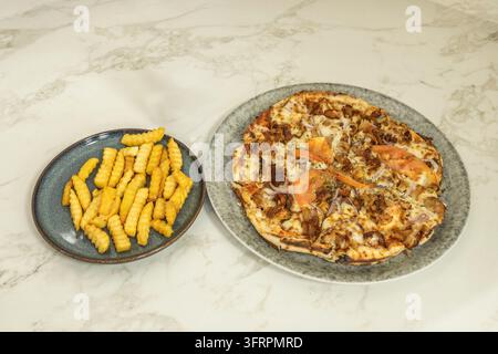 Pizza au poulet garnie de tranches de tomate accompagnée de frites ondulées servies sur des assiettes en céramique sur une table en marbre. Banque D'Images