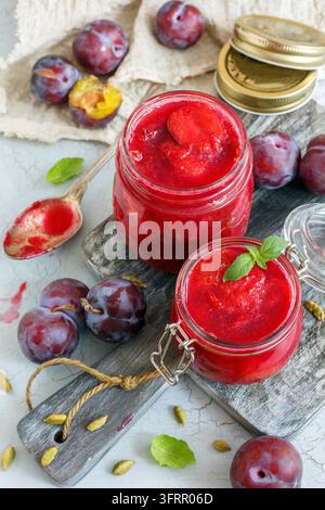 Bocaux en verre de confiture de prunes maison au cardamome et prunes dans un vieux bol en bois sur un fond en béton texturé Banque D'Images