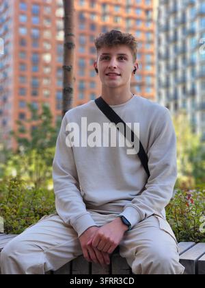 Jeune homme portant une chemise blanche et un pantalon kaki est assis sur un banc. Il porte un sac à dos noir et une montre Banque D'Images