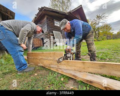 Deux hommes travaillent sur un projet dans une cour. Un homme utilise une tronçonneuse pour couper un morceau de bois Banque D'Images