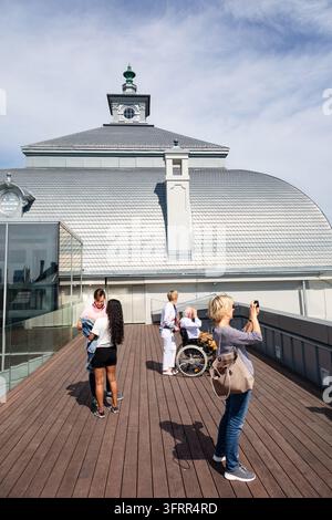 TOURISTES SUR UNE TERRASSE D'OBSERVATION SUR LE TOIT, RÉPORTAGE DE VOYAGE À RIGA, RIGA, LETTONIE : Le Musée National d'Art de Lettonie. Un groupe de personnes, certaines prenant des photos avec un téléphone portable et un autre poussant une personne en fauteuil roulant, se tient sur une plateforme d'observation en bois devant un bâtiment au toit distinctif. INFO : Riga, la capitale de la Lettonie, est la plus grande ville des États baltes. Mélant histoire médiévale et charme cosmopolite, elle est mondialement reconnue pour son architecture Art Nouveau exceptionnelle inscrite au patrimoine mondial de l'UNESCO et sa vieille ville pavée magnifiquement préservée. Banque D'Images