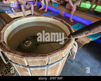 Seau en bois avec une cuillère dedans. Le seau est rempli d'eau. Il y a des gens assis autour du seau dans un bain à vapeur de sauna Banque D'Images
