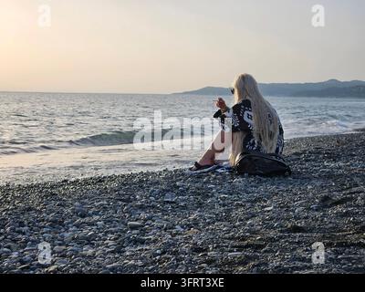 Femme aux longs cheveux blonds et une cigarette assise sur une plage. Elle porte une chemise noire et blanche et a un sac à dos sur ses genoux Banque D'Images