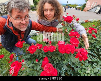 Un homme et une femme se tiennent devant un buisson de roses rouges. L'homme porte des lunettes et la femme porte une veste. Ils regardent tous les deux la r Banque D'Images