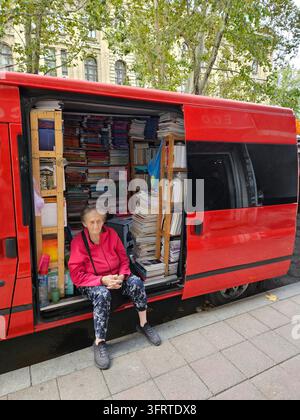 BELGRADE - 25 SEPT 2025 : Femme assise à l'arrière d'une camionnette rouge. La camionnette est pleine de livres et possède une étagère à l'arrière. La femme porte un sweat à capuche rouge Banque D'Images