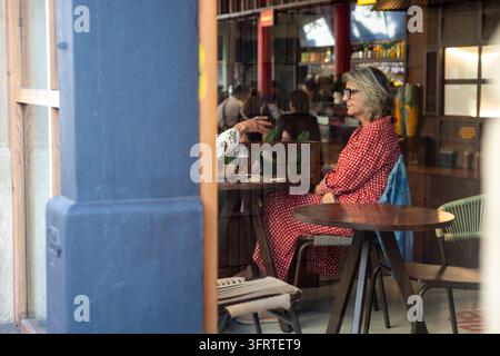Barcelone, Espagne, 13 août 2026 – Une femme âgée vêtue d'une robe à motifs rouges est assise à une table de café, vue à travers une fenêtre, tandis qu'une autre personne gesticule Banque D'Images