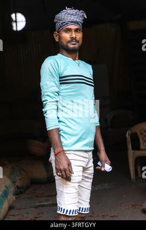 Portrait d'un jeune ouvrier tenant un petit papier dans sa main avec un short blanc et des sacs empilés dans l'espace sombre derrière lui, Bengale occidentale, Howrah, Inde Banque D'Images