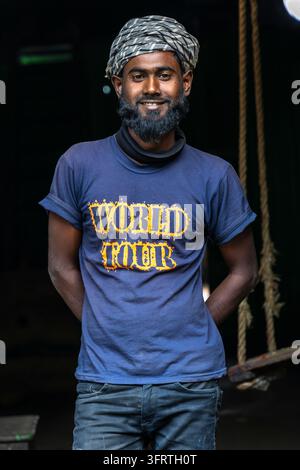 Portrait d'un jeune travailleur amical avec une écharpe noire au cou et un jean sombre, debout dans un entrepôt sombre avec des cordes suspendues en arrière-plan, Bengale occidentale Banque D'Images