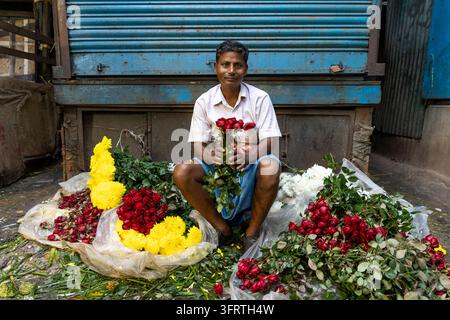 Un homme souriant en chemise blanche et lungi est assis par terre tenant quelques roses entourées de roses rouges et de soucis jaunes au marché aux fleurs, Wes Banque D'Images