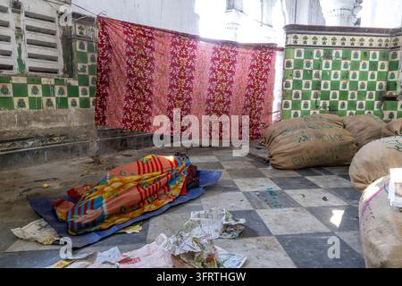 Une couverture imprimée pliée repose sur un sol en marbre noir et blanc dans un hall usé à côté de sacs en toile de jute et d'un tissu rouge accroché à un mur carrelé vert au f Banque D'Images