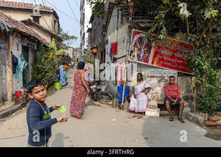 Rue de quartier avec un garçon tenant un jouet à ficelle, une femme en robe rouge marchant et une femme et un homme assis près d'un panneau de sanctuaire peint, Bengale occidentale, K Banque D'Images