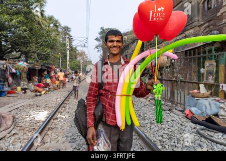 Vendeur joyeux avec un sac à dos et un paquet de cœurs rouges et de ballons torsadés longs se tient en file près des familles assises près de leurs abris, West Banque D'Images