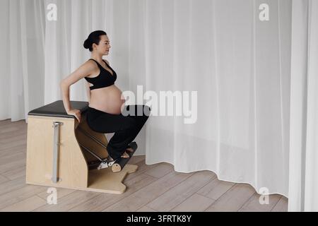 Femme enceinte assise sur une boîte de pilates en bois dans une pièce studio minimaliste et lumineuse Banque D'Images