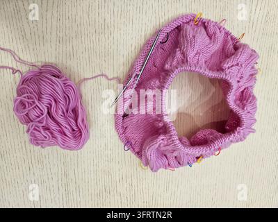 Une boule de laine rose repose à côté du projet de tricot. L'anneau tricoté présente des motifs de mailles colorés. L'aiguille est insérée dans le travail, les épingles maintiennent la forme. Le tissu texturé montre le processus de création artisanale en cours Banque D'Images