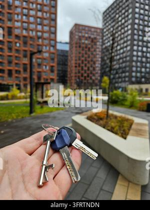Une main tient des clés près de bâtiments modernes et de verdure. Cadre urbain avec des structures hautes et des allées pavées. Les clés suggèrent une nouvelle maison ou un nouvel appartement. L'arrière-plan comprend des arbres, des bacs à plantes Banque D'Images
