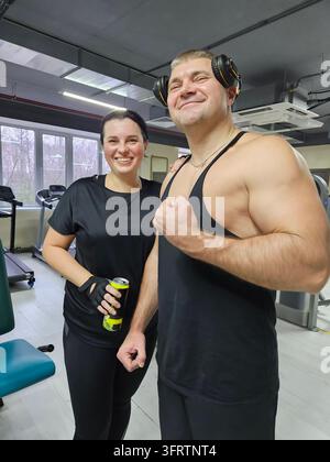 Deux personnes sourient dans un cadre de salle de sport. L'une porte des écouteurs et un débardeur. L'autre tient une canette et porte une tenue noire. Ils se tiennent près d'équipements de gymnastique dans une pièce bien éclairée Banque D'Images