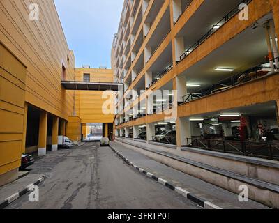 Garage de stationnement à plusieurs niveaux avec murs en béton jaune. Des voitures garées sur plusieurs étages sous des lumières au plafond. Une route d'accès passe entre les structures adjacentes des bâtiments Banque D'Images