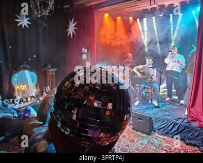 Le globe disco domine l'avant-plan, reflétant les lumières de scène. Un groupe joue sur une scène faiblement éclairée. Les projecteurs illuminent les musiciens et les instruments. Des décorations en forme d'étoile pendent au-dessus de la scène Banque D'Images