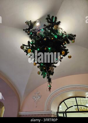 Un sapin de Noël en forme de lustre est suspendu à un plafond voûté. Des lumières vertes illuminent des ornements dorés. Un arbre décoratif suspendu au-dessus d'une porte voûtée. Des murs rose doux encadrent l'installation festive Banque D'Images