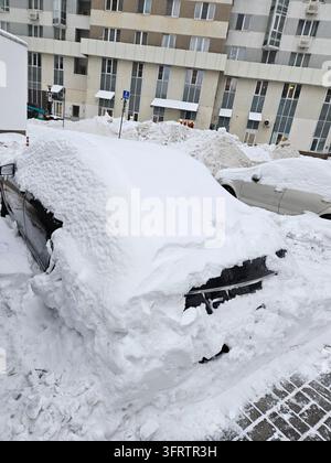 Voiture enneigée garée près d'un immeuble d'habitation. D'épais tas de neige sur le toit et les côtés du véhicule. Cadre urbain avec plusieurs voitures garées. Bâtiment doté de balcons et de fenêtres Banque D'Images