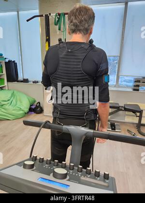 Un homme se tient sur un gilet d'entraînement de résistance sur tapis de course. L'équipement ajuste les paramètres sur le panneau de contrôle. L'équipement de salle de gym est visible. La posture de l'homme suggère une séance d'entraînement active avec stimulation électromusculaire Banque D'Images