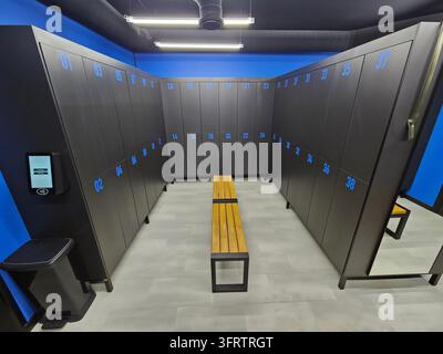 Vestiaire moderne avec armoires éclairées en bleu et banc en bois. Design épuré et propre avec murs et sol noirs. Distributeur de désinfectant pour les mains et poubelle visibles Banque D'Images