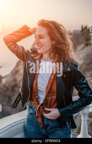 Femme coucher de soleil côte jeune femme souriante portant une veste en cuir noir posant sur un balcon avec vue sur la mer à l'heure dorée Banque D'Images