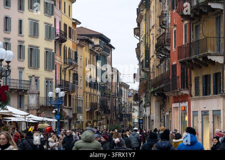 Rue piétonne animée bordée de bâtiments historiques colorés, de balcons, de boutiques, d'étals de marché et de visiteurs hivernaux dans le centre de Vérone. Banque D'Images