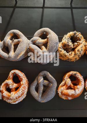 Bretzels artisanaux suspendus à un mur à chevilles sombre dans une vitrine de boulangerie, présentant des variétés semées, salées et recouvertes de pavot. Tons dorés chauds, texture rustique et Banque D'Images