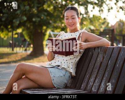 Une femme asiatique d'âge moyen heureuse se détend dans un parc en été, elle lit un livre. Banque D'Images