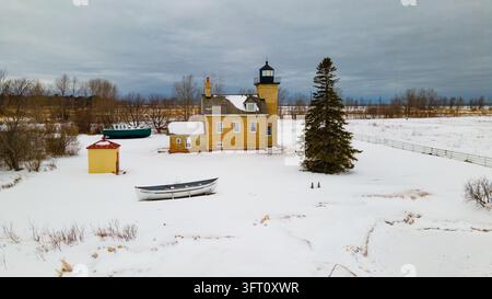 Vue aérienne du phare d'Ontonagon entouré de neige avec un petit bateau sur le sol gelé, un grand arbre à feuilles persistantes et un ciel couvert à Onton Banque D'Images