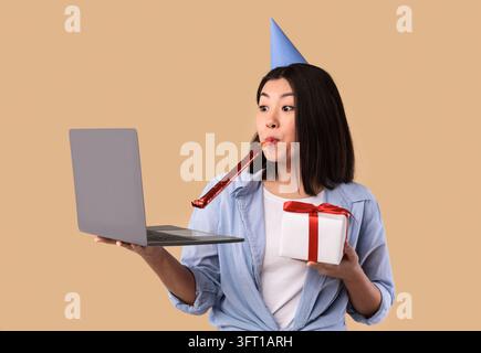 Concept de salutations en ligne. Drôle jeune femme asiatique portant chapeau, tuyau de corne de fête soufflant, tenant l'ordinateur portable et boîte-cadeau enveloppé avec cadeau faire en ligne Banque D'Images