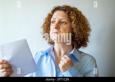 Femme d'âge moyen pensive tenant un document et ajustant son col dans un portrait neutre Banque D'Images