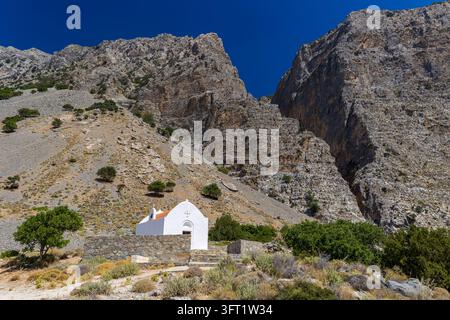 Chapelle aux murs blancs avec tuiles rouges sur un flanc de montagne aride en Grèce, mur de clôture bas en pierre à l'entrée d'une gorge rocheuse Banque D'Images