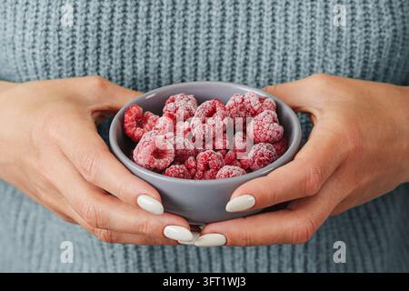 Grossissement de framboises givées dans un petit bol gris tenu à deux mains. Arrière-plan de baies congelées pour les desserts froids, l'alimentation saine et le stockage des fruits. Banque D'Images