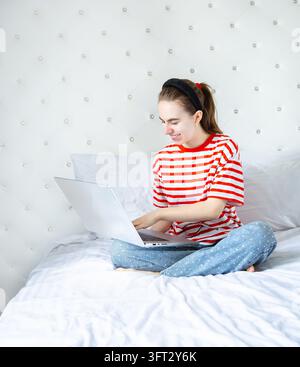 Jeune femme heureuse portant une chemise rayée et utilisant un ordinateur portable, assise confortablement sur un lit blanc à la maison. Banque D'Images