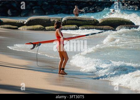 Femme tenant une longue planche de surf au rivage avec des vagues se brisant contre un brise-lames rocheux sous un soleil éclatant et vue sur la plage de sable, 35 Banque D'Images