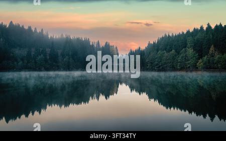 Lac Karagol à Savsat, Artvin, Turquie, à l'aube avec un ciel rose et orange, brouillard sur l'eau et forêt de sapins sombres reflétés sur la surface calme Banque D'Images