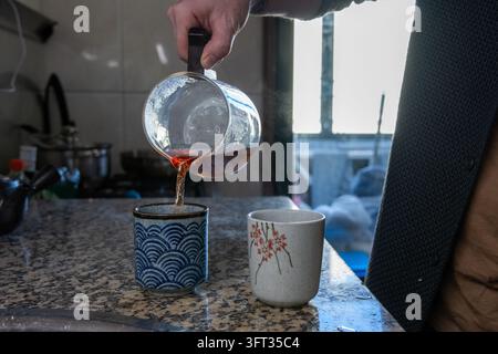 Cliché d'action des mains d'un homme versant du thé chaud fraîchement infusé d'une carafe en verre transparent dans une tasse à thé en céramique décorée sur un comptoir de cuisine Banque D'Images