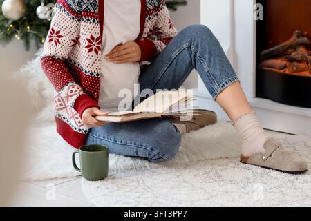 Confort de la grossesse et de la maternité, femme enceinte lisant un livre au coin du feu pendant les vacances d'hiver dans un cadre domestique confortable avec une boisson chaude Banque D'Images