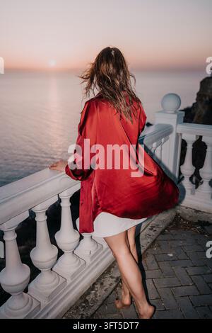 Femme coucher de soleil océan jeune femme en chemise de soie rouge regardant la mer depuis un balcon blanc lors d'une soirée paisible Banque D'Images